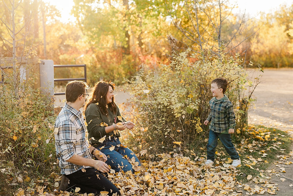 A family playing in autumn leaves, smiling and laughing. The setting is a sunlit park with trees in vibrant fall colors.