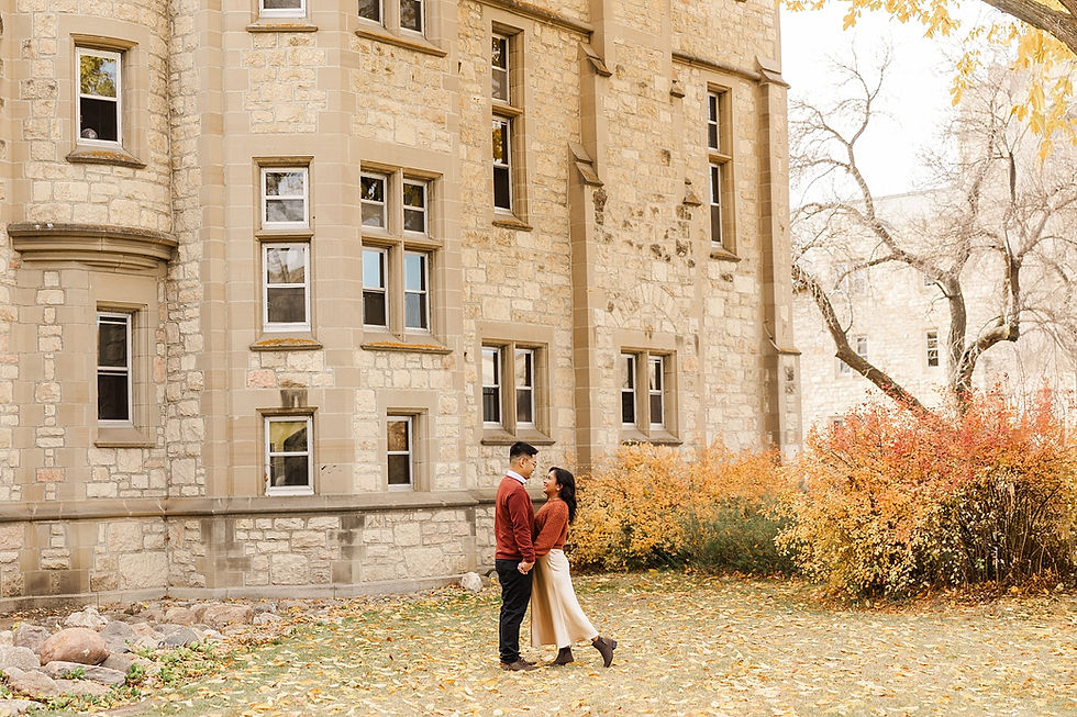Couple smiling and holding hands near a stone building, surrounded by autumn leaves and colorful bushes, conveying a warm, romantic mood.