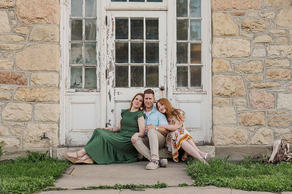 Three people sit together on a stone doorstep. The man is in the middle, with women on each side. Background features an old wooden door.