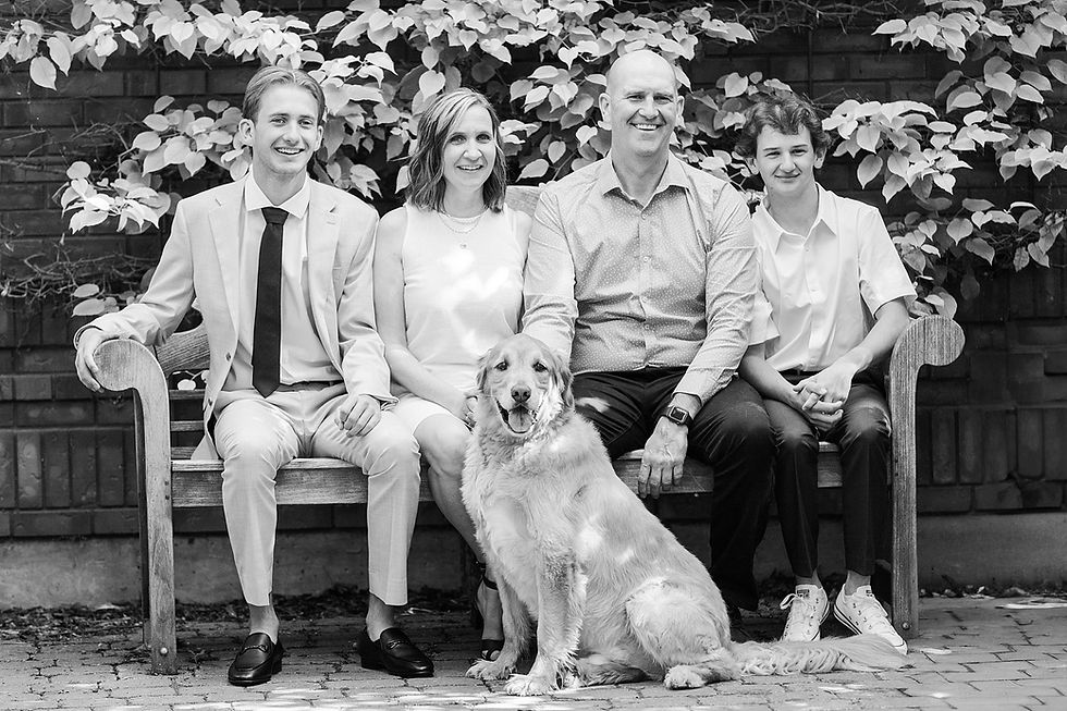 A family of four and a dog sit on a bench outdoors against leafy vines. They are smiling, creating a warm, joyful mood. Black and white image.