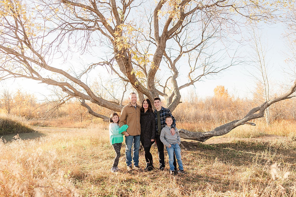Family of five smiles under a large, leafless tree in an autumn field. Bright, sunny day with golden grasses and clear blue sky.
