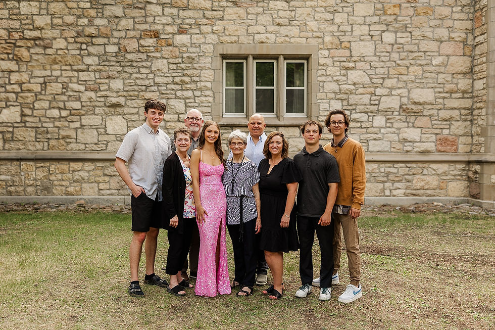 A group of nine people smiles in front of a stone building. A woman in a pink dress stands mid-front, with a casual outdoor setting.