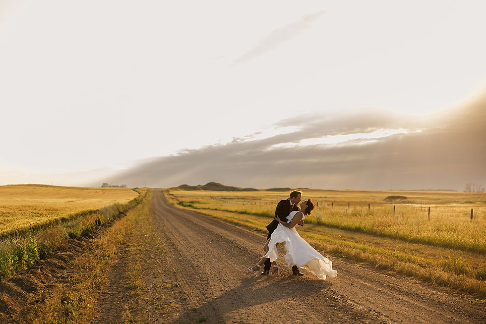 A couple dances on a rural dirt road at sunset. The landscape is wide and golden, creating a romantic, serene atmosphere.