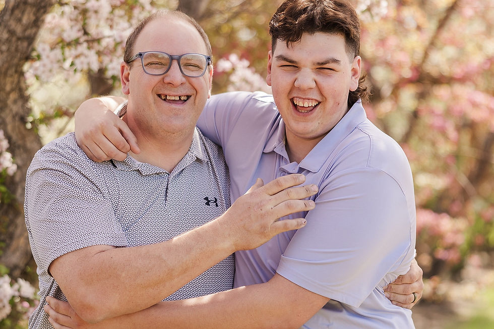 Two people hug and smile outdoors, surrounded by blooming pink flowers. One wears a patterned shirt and glasses, the other a light purple shirt.