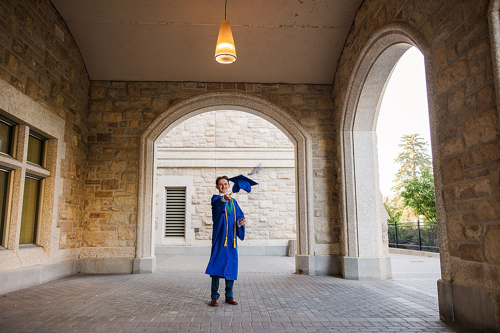 Young graduate in blue gown tosses cap under stone archway, smiling in a sunny courtyard.