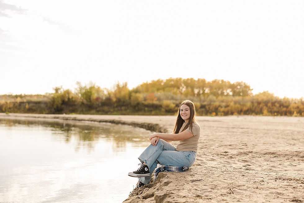 A young woman sits smiling on a sandy lakeside, wearing jeans and sneakers. Autumn trees are in the sunny background, reflecting calm serenity.