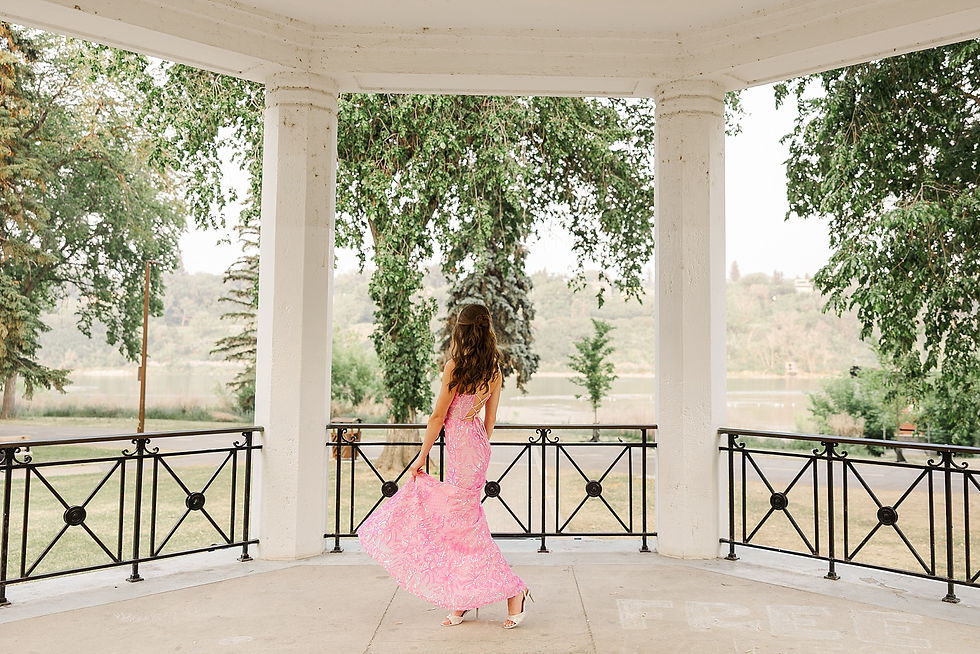 Woman in pink dress stands in a gazebo, surrounded by greenery, overlooking a river. She gracefully holds her dress, creating a serene mood.