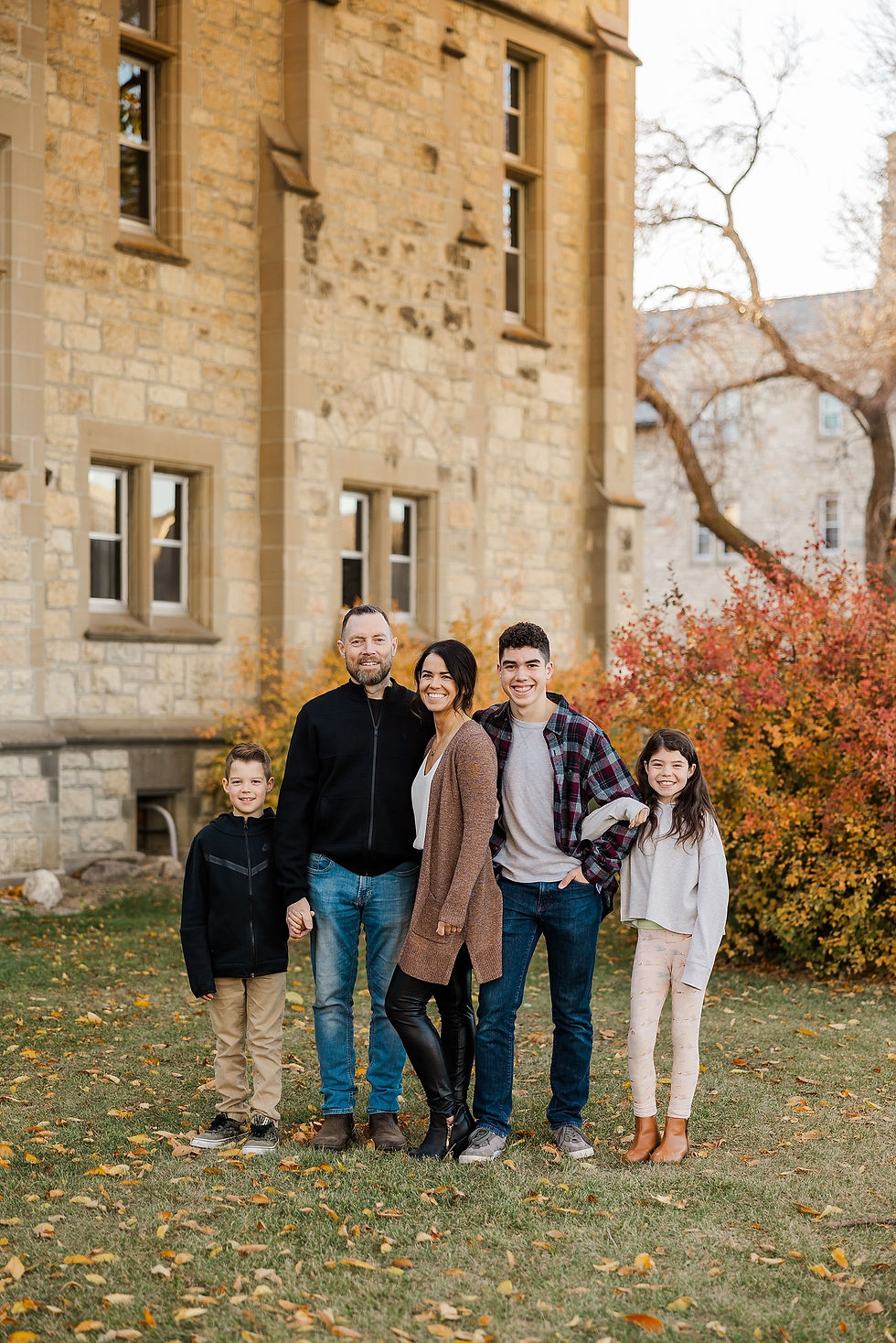 Family of five smiling in front of a stone building, with autumn foliage and grass. Everyone looks happy and relaxed, dressed casually.