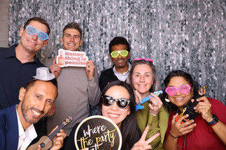 Group of coworkers wearing playful sunglasses and holding party signs while posing in a corporate event photo booth setup.
