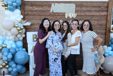 Group of five women posing at a baby shower photo booth in front of a wooden backdrop with blue and white balloon decor and the name Archer displayed above.