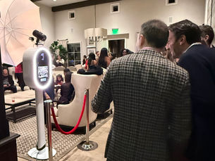People gather near a photo booth in a stylish lounge. Red carpet and velvet rope add a formal touch. Mood is lively and social.