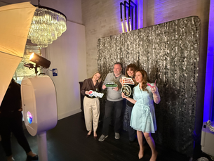 Four people smiling, holding signs at a photo booth with a sparkly backdrop. A chandelier and purple lighting decorate the room.