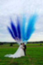 Bride and groom standing together in a green field with vibrant blue and purple smoke rising behind them.