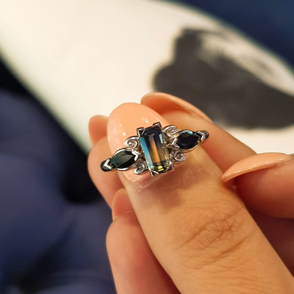 Hand holding a white gold ring with an elongated green-blue parti color sapphire. Background is blurred dark blue and white. Nails are polished in peach.
