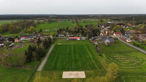 Aerial view of a small German village with farmhouses, a football pitch, green fields, and gardens stretching into the countryside.
