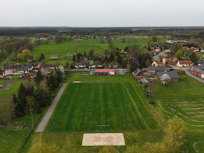 Aerial view of a small German village with farmhouses, a football pitch, green fields, and gardens stretching into the countryside.