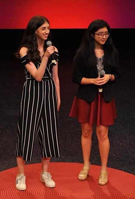 Me and my VP Marianne stand on a black box stage. The ground is covered in a red circular rug and the back wall of the stage is lit with red lighting. Stephanie is smiling and holding a microphone. 