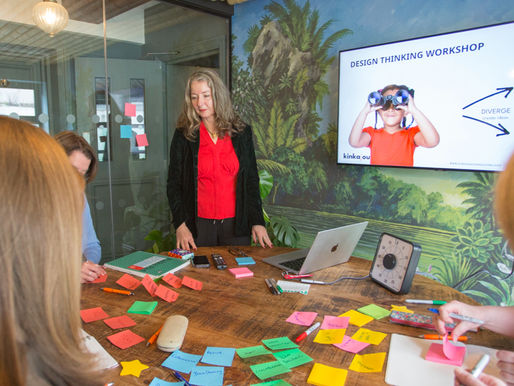 Woman leading a Design Thinking workshop, surrounded by colorful sticky notes on a table. A presentation screen displays text and an image.