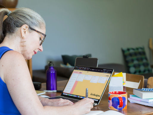 Woman in blue top writes in a notebook at a wooden table with a laptop displaying "kinkajou," colorful mug, books, and purple bottle nearby.
