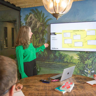 Woman in green shirt presenting coaching timeline on screen. Two people seated, with notebooks and a laptop on the wooden table. Jungle mural.