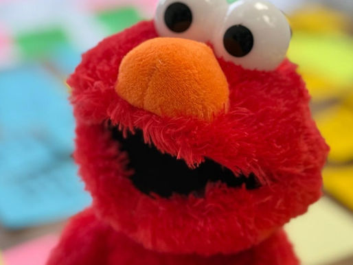 EMLO red puppet with large eyes and an orange nose smiles in a colorful background of scattered post-it notes, creating a playful mood.