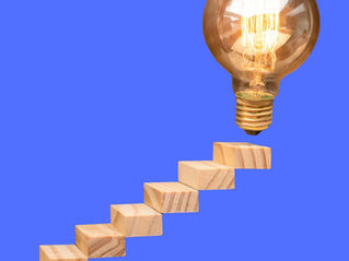 Wooden blocks form steps leading to a glowing lightbulb against a blue background, symbolizing ideas and progress