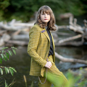A person in a yellow plaid suit stands near a river with driftwood. Their expression is neutral, and greenery is in the background.