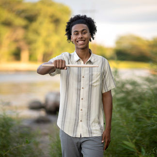 Person by a river, wearing a striped shirt and jeans. Background shows trees. Calm and relaxed mood.