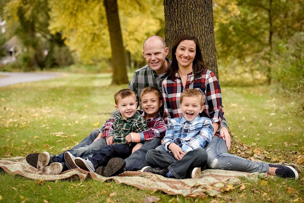 Family of five in plaid shirts sit smiling on a blanket under a tree in a green park. Leaves are scattered on the grass; warm, joyful mood.