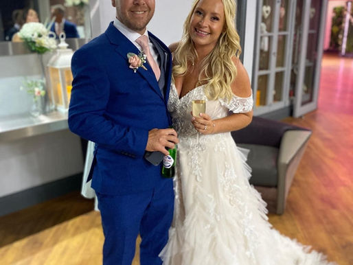 Real bride Lucy, wearing a textured lace and tulle Mori Lee wedding gown with off the shoulder sleeves, smiles next to her groom Chris who is wearing a vibrant blue suit with a blush pink tie and rose buttonhole.