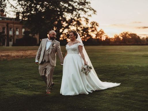 Real bride Jade wearing a classic white A-line Essense of Australia wedding dress with an embellished off-the-shoulder bodice holds hands with her groom Jody, who wears a tan three-piece suit, as they walk across the lawns of Braxted Park at sunset.