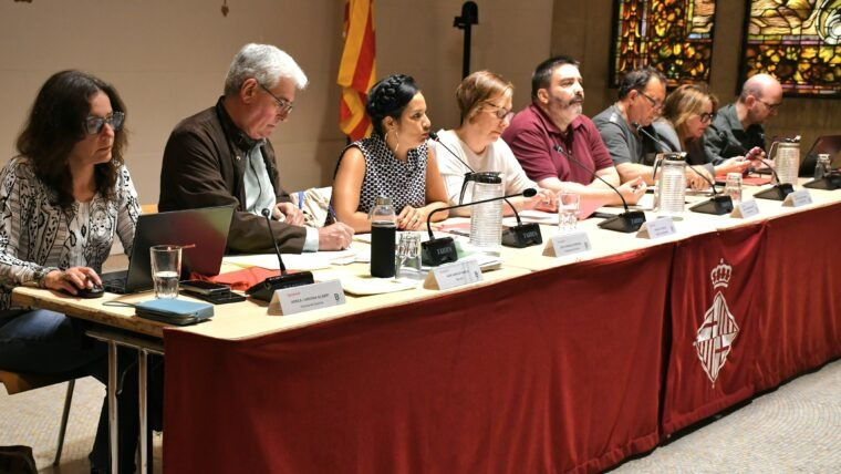 Set persones en una taula de reunió, prenent notes i parlant en una sala amb vitralls i banderes, amb estovalló vermell