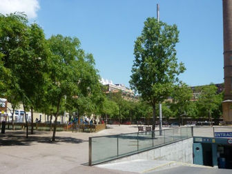 Plaça amb arbres verds i una xemeneia alta al fons. Senyal d'entrada visible. Ambient assolellat i tranquil. Gent passejant a prop.