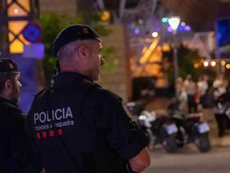 Policies patrullant de nit; uniformes negres amb "Policia" visible. Fons amb llums i grups de gent. Ambient de vigilància urbana.