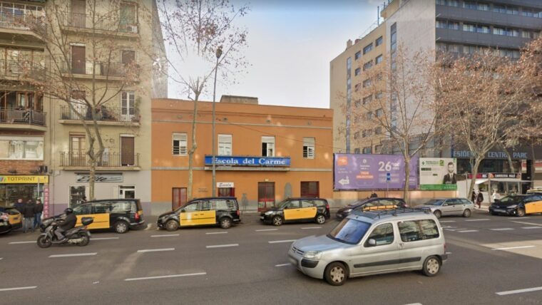 Carrer urbà amb cotxes i motos davant l’Escola del Carme i edificis al fons; ambient assolellat i tranquil.