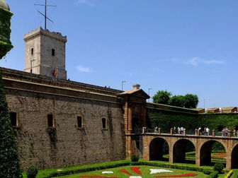 Fortalesa amb murs de pedra coberts d'heura, persones passejant per un pont sobre jardins acolorits. Cel blau clar i tranquil.
