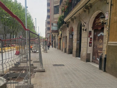 Carrer amb vorera tancada per obres. Edificis antics a la dreta, tanques i arbres a l'esquerra. Una persona camina en la distància.