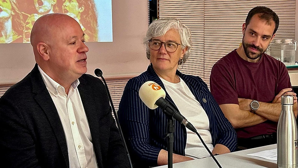 Tres persones assegudes en una conferència. Un home parla al micròfon. Fons de persianes i pantalla. Ambient professional i atent.