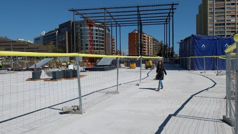 Una dona camina en una zona en construcció, envoltada de tanques de seguretat. Fons de gratacels i estructures metàl·liques sota un cel blau.