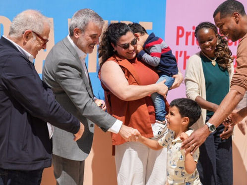 Un grup de persones somrients dona la mà a un nen petit. El fons és colorit amb text parcialment visible. L'ambient és alegre.