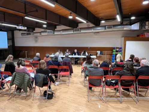 Persones assegudes en cadires vermelles escoltant una taula rodona en una sala amb sostres de fusta. Projector i càmera a l'esquerra. Ambient seriós.