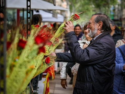 Home d'edat avançada somrient, agafant roses envoltades en tela groga en un mercat a l'aire lliure. Gent al voltant amb expressions alegres.