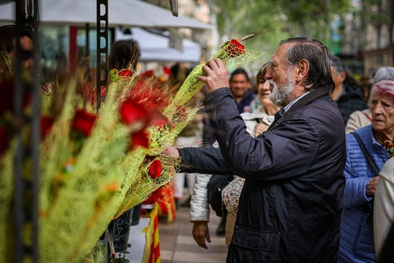 Home d'edat avançada somrient, agafant roses envoltades en tela groga en un mercat a l'aire lliure. Gent al voltant amb expressions alegres.