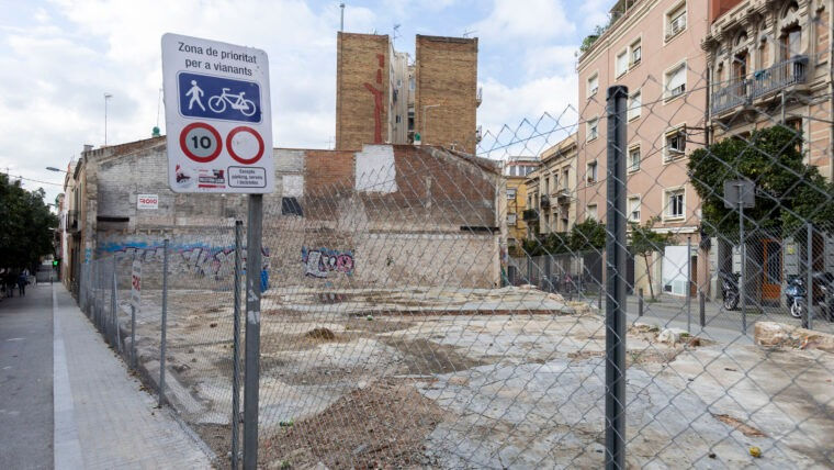 Solar buit amb tanques metàl·liques. Senyal de trànsit que indica prioritat per a vianants i ciclistes. Edificis i arbres al fons.