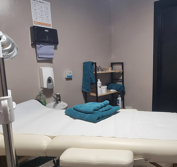 Interior photography of a clean treatment room. A massage table with folded towels is central, with shelves stocked with products, suggesting a professional and relaxing environment.