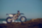 A dirt bike stands on rocky terrain by the ocean. The bike is mostly silver with black and yellow details, set against a clear blue sky.