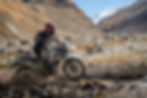 A motorcyclist in red helmet rides through a rocky stream, splashing water, in a barren mountain valley under a clear blue sky.
