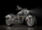 Silver Harley-Davidson motorcycle with a shining headlight in a dark studio setting. Polished chrome details, sleek design, and an emblem on the tank.