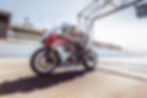 Motorcyclist on a red and white Suzuki racing bike speeds on a sunlit track, stadium backdrop, wearing racing gear, text "Suzuki" visible.