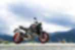 Motorcycle with black body and red wheels on a scenic mountain road under cloudy skies, evoking a sense of adventure and freedom.
